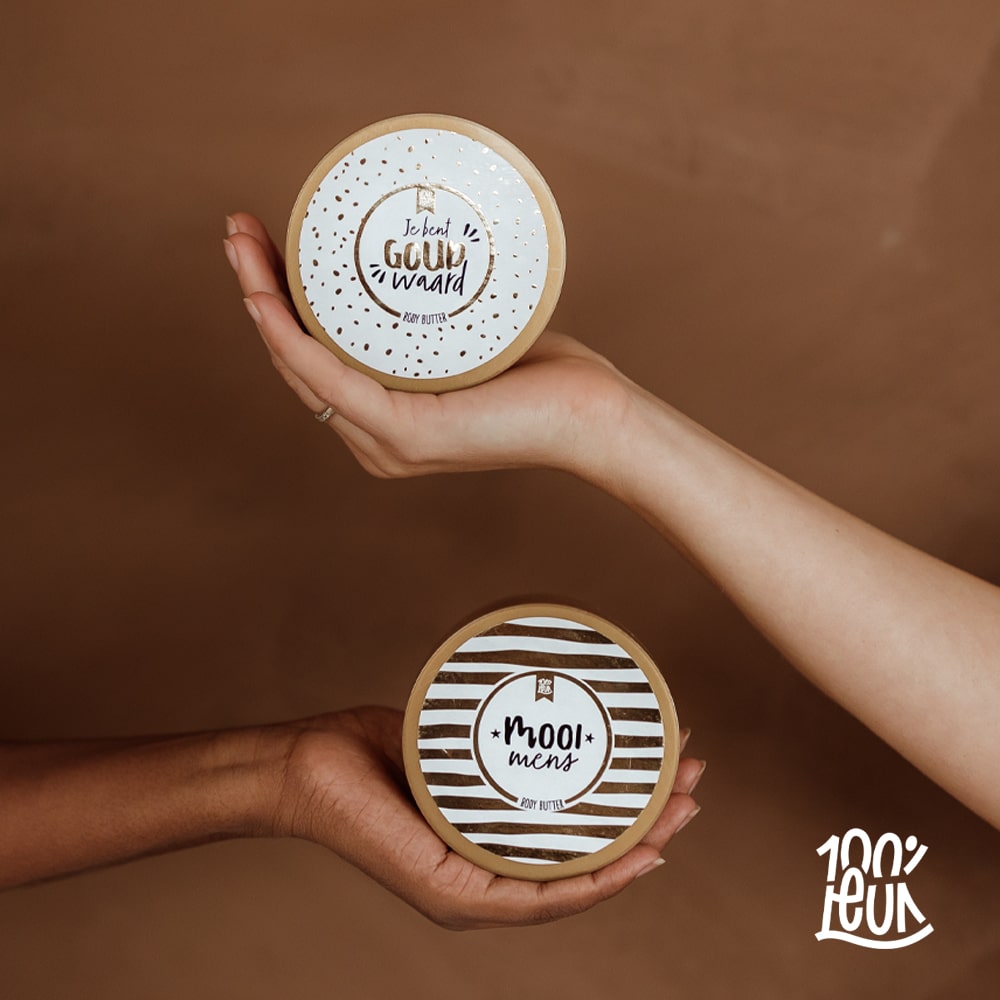 Twee potten bodybutter met de tekst 'je bent goud waard' en 'mooi mens', vastgehoude op twee handen