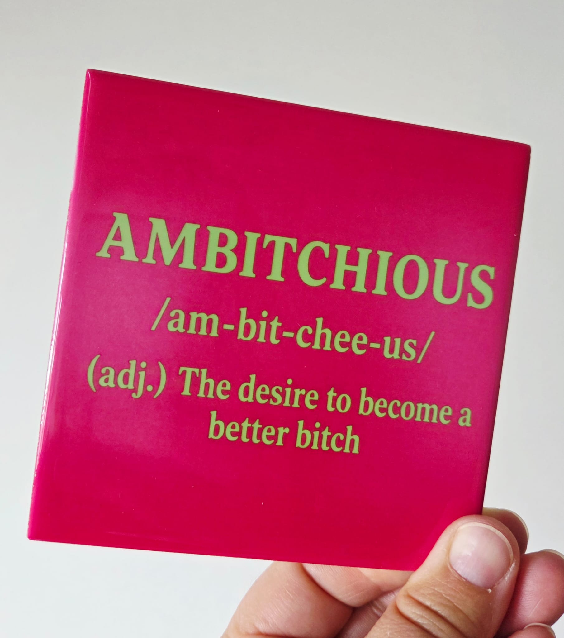 Roze tegel met groene letters en de tekst Ambitchious. The desire to become a better bitch